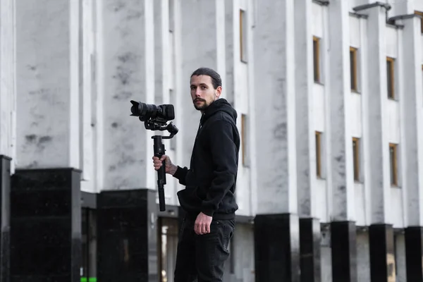 Jovem videógrafo profissional segurando câmera profissional no estabilizador de 3 eixos gimbal. Pro equipamento ajuda a fazer vídeo de alta qualidade sem tremer. Cameraman vestindo capuz preto fazendo um vídeo . — Fotografia de Stock