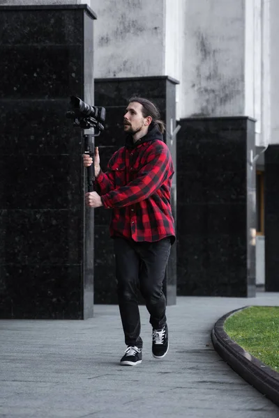 Junger professioneller Videofilmer mit professioneller Kamera auf 3-Achsen-Gimbal-Stabilisator. Pro-Ausrüstung hilft, qualitativ hochwertige Videos ohne Schütteln zu machen. Kameramann im roten Hemd dreht ein Video. — Stockfoto