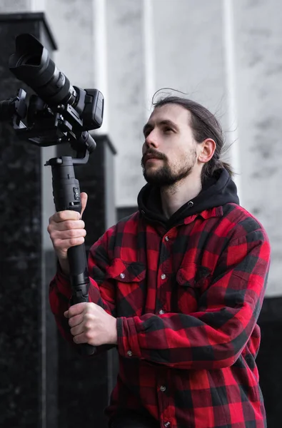 Ung professionell videograf som håller professionell kamera på 3-axlig gimbal stabilisator. Pro-utrustning hjälper till att göra högkvalitativ video utan att skaka. Kameraman bär röd skjorta gör en video. — Stockfoto