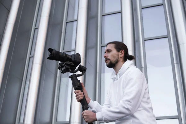 Jovem videógrafo profissional segurando câmera profissional no estabilizador de 3 eixos gimbal. Pro equipamento ajuda a fazer vídeo de alta qualidade sem tremer. Cameraman vestindo capuz branco fazendo um vídeo . — Fotografia de Stock