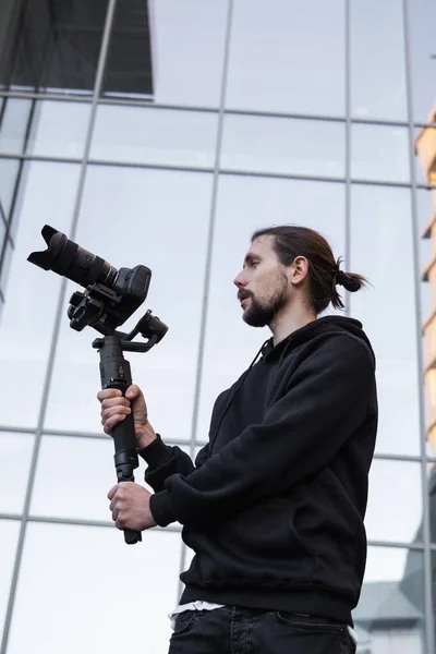 Young Professional videographer holding professional camera on 3-axis gimbal stabilizer. Pro equipment helps to make high quality video without shaking. Cameraman wearing white hoodie making a videos.