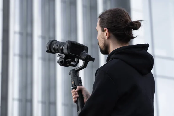 Young Professional videographer holding professional camera on 3-axis gimbal stabilizer. Pro equipment helps to make high quality video without shaking. Cameraman wearing white hoodie making a videos.