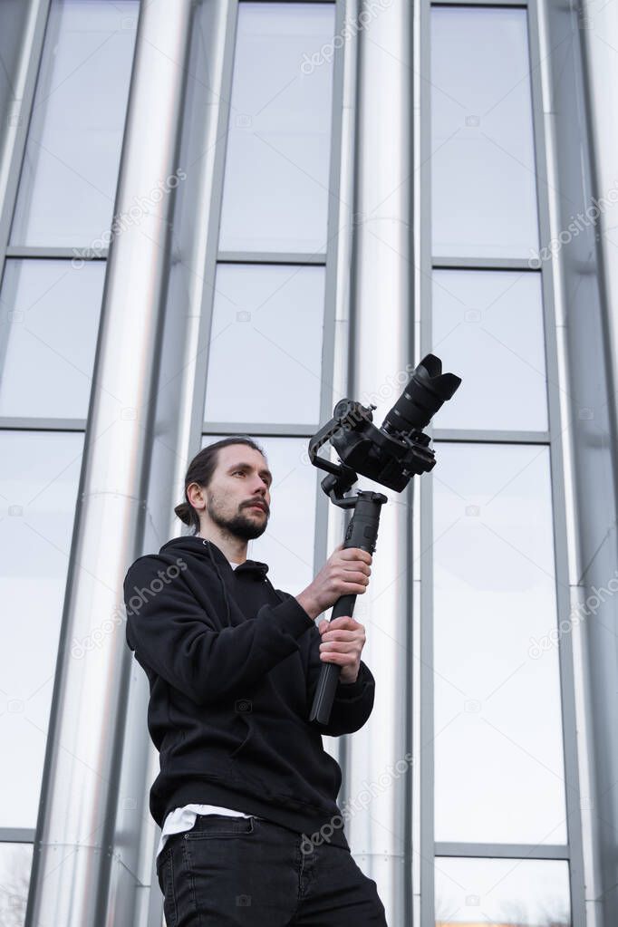 Young Professional videographer holding professional camera on 3-axis gimbal stabilizer. Pro equipment helps to make high quality video without shaking. Cameraman wearing white hoodie making a videos.