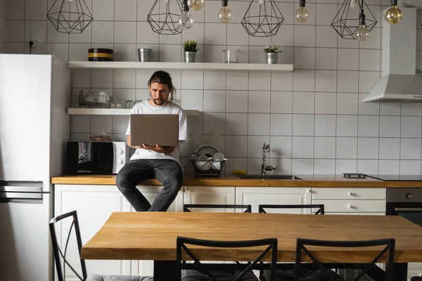 Ελεύθερος επαγγελματίας που εργάζεται από το σπίτι κάθεται σε έναν πάγκο κουζίνας και χρησιμοποιώντας το lap-top. Γενειοφόρος που δουλεύει με λάπτοπ και διαβάζει ειδήσεις. Όμορφος επιτυχημένος επιχειρηματίας που εργάζεται στο σύγχρονο σπίτι του. — Φωτογραφία Αρχείου