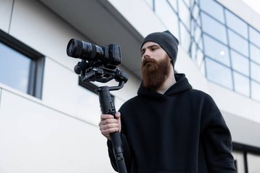 Siyah kapüşonlu sakallı profesyonel kameraman elinde 3 eksenli titreşim dengeleyicisi var. Film yapımcısı profesyonel bir sinema kamerasıyla harika bir video çekiyor. Görüntü Yönetmeni.