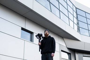 Siyah kapüşonlu sakallı profesyonel kameraman elinde 3 eksenli titreşim dengeleyicisi var. Film yapımcısı profesyonel bir sinema kamerasıyla harika bir video çekiyor. Görüntü Yönetmeni.