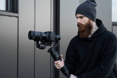 Siyah kapüşonlu sakallı profesyonel kameraman elinde 3 eksenli titreşim dengeleyicisi var. Film yapımcısı profesyonel bir sinema kamerasıyla harika bir video çekiyor. Görüntü Yönetmeni.