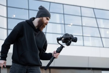 Siyah kapüşonlu sakallı profesyonel kameraman elinde 3 eksenli titreşim dengeleyicisi var. Film yapımcısı profesyonel bir sinema kamerasıyla harika bir video çekiyor. Görüntü Yönetmeni.