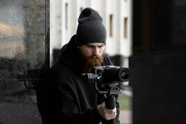 Vidéographe professionnel barbu en sweat à capuche noir tenant une caméra professionnelle sur stabilisateur de cardan 3 axes. Cinéaste faire une grande vidéo avec une caméra de cinéma professionnel. Directeur de la photographie . — Photo