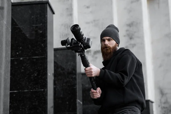 Bearded Professional videographer in black hoodie holding professional camera on 3-axis gimbal stabilizer. Filmmaker making a great video with a professional cinema camera. Cinematographer. — Stock Photo, Image