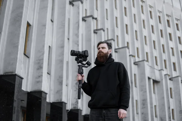 Videografo professionale barbuto con cappuccio nero con fotocamera professionale su stabilizzatore gimbale a 3 assi. Filmmaker fare un grande video con una macchina fotografica cinematografica professionale. Cinematografo . — Foto Stock