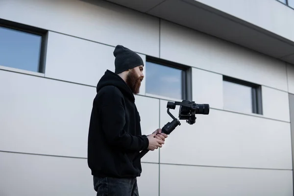 Vídeo profissional barbudo com capuz preto segurando câmera profissional no estabilizador de 3 eixos gimbal. Filmmaker fazendo um grande vídeo com uma câmera de cinema profissional. Fotografo . — Fotografia de Stock