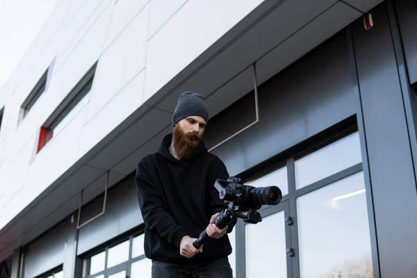 Videografo professionale barbuto con cappuccio nero con fotocamera professionale su stabilizzatore gimbale a 3 assi. Filmmaker fare un grande video con una macchina fotografica cinematografica professionale. Cinematografo . — Foto Stock