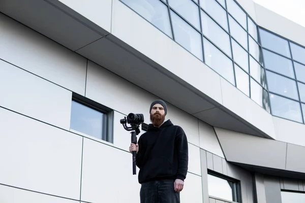 Videografo professionale barbuto con cappuccio nero con fotocamera professionale su stabilizzatore gimbale a 3 assi. Filmmaker fare un grande video con una macchina fotografica cinematografica professionale. Cinematografo . — Foto Stock