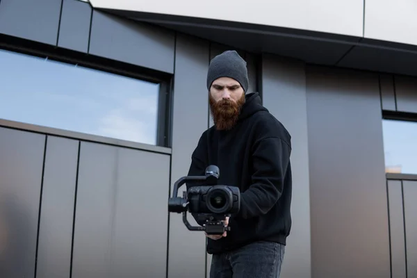 Γενειάδα επαγγελματική videographer σε μαύρο hoodie κρατώντας επαγγελματική κάμερα σε 3-άξονα gimbal σταθεροποιητή. Κινηματογραφιστής κάνει ένα μεγάλο βίντεο με μια επαγγελματική κάμερα κινηματογράφου. Κινηματογράφος. — Φωτογραφία Αρχείου