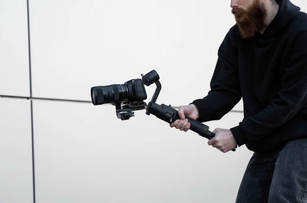 Vousatý profesionální kameraman v černé mikině s profesionální kamerou na tříosém gimbálním stabilizátoru. Filmař natáčí skvělé video s profesionální kamerou. Kameraman. — Stock fotografie