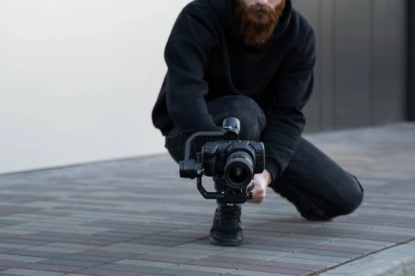 Vídeo profissional barbudo com capuz preto segurando câmera profissional no estabilizador de 3 eixos gimbal. Filmmaker fazendo um grande vídeo com uma câmera de cinema profissional. Fotografo . — Fotografia de Stock