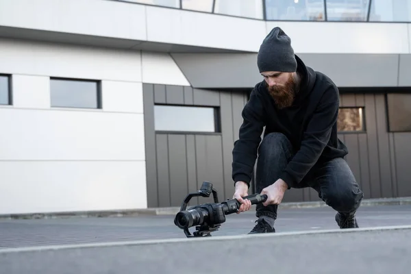 Videografo professionale barbuto con cappuccio nero con fotocamera professionale su stabilizzatore gimbale a 3 assi. Filmmaker fare un grande video con una macchina fotografica cinematografica professionale. Cinematografo . — Foto Stock