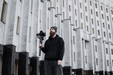 Siyah kapüşonlu sakallı profesyonel kameraman elinde 3 eksenli titreşim dengeleyicisi var. Film yapımcısı profesyonel bir sinema kamerasıyla harika bir video çekiyor. Görüntü Yönetmeni.