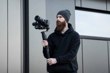 Siyah kapüşonlu sakallı profesyonel kameraman elinde 3 eksenli titreşim dengeleyicisi var. Film yapımcısı profesyonel bir sinema kamerasıyla harika bir video çekiyor. Görüntü Yönetmeni.