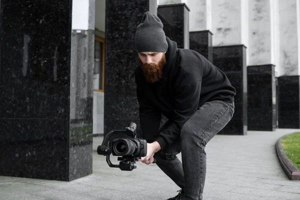 Бородатий професійний відеооператор в чорному светрі тримає професійну камеру на 3-осьовому стабілізаторі. Кінематографіст знімає чудове відео з професійною кінокамерою. Кінематографіст . — стокове фото