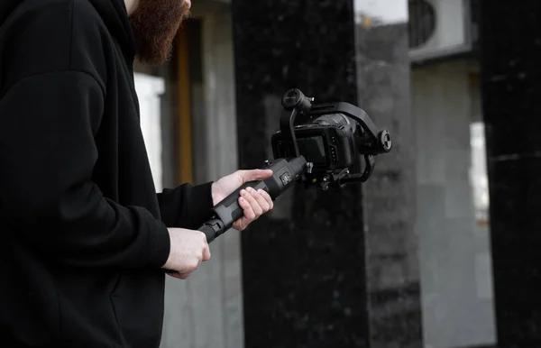 Videografo professionale barbuto con cappuccio nero con fotocamera professionale su stabilizzatore gimbale a 3 assi. Filmmaker fare un grande video con una macchina fotografica cinematografica professionale. Cinematografo . — Foto Stock