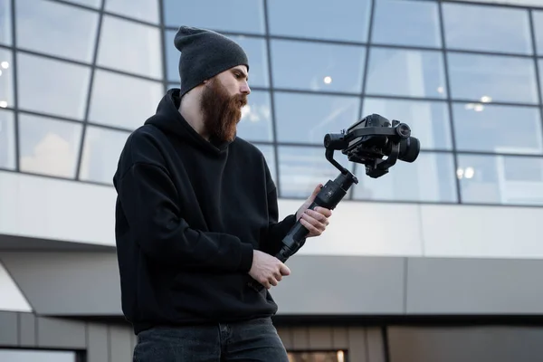 Videografo professionale barbuto con cappuccio nero con fotocamera professionale su stabilizzatore gimbale a 3 assi. Filmmaker fare un grande video con una macchina fotografica cinematografica professionale. Cinematografo . — Foto Stock