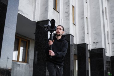 3 eksenli titreşimli dengeleyicide profesyonel kamera tutan genç profesyonel kameraman. Pro ekipmanlar titremeden yüksek kaliteli video çekmeye yardımcı olur. Siyah kapüşonlu bir kameraman video çekiyor..