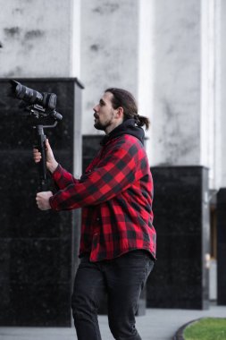 Young Professional videographer holding professional camera on 3-axis gimbal stabilizer. Pro equipment helps to make high quality video without shaking. Cameraman wearing red shirt making a videos. clipart