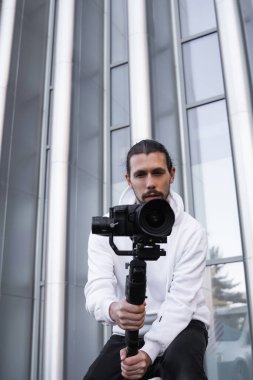 3 eksenli titreşimli dengeleyicide profesyonel kamera tutan genç profesyonel kameraman. Pro ekipmanlar titremeden yüksek kaliteli video çekmeye yardımcı olur. Beyaz kapüşonlu bir kameraman video çekiyor..