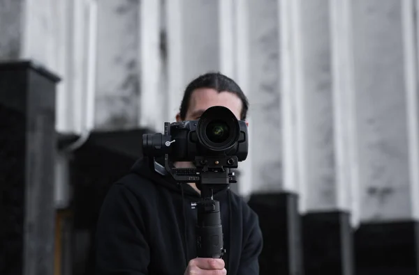 Young Professional videographer holding professional camera on 3-axis gimbal stabilizer. Pro equipment helps to make high quality video without shaking. Cameraman wearing black hoodie making a videos.