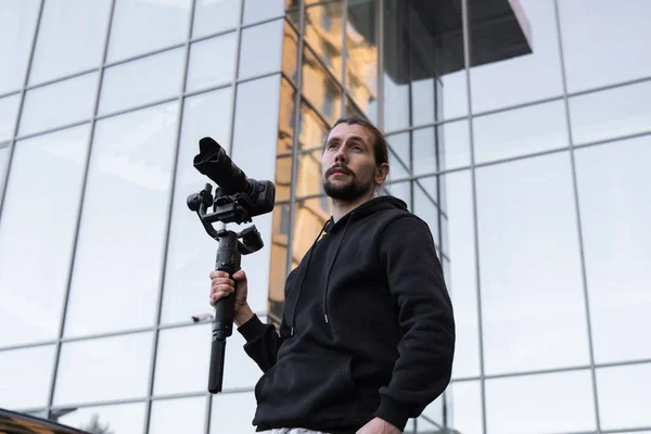 Jovem videógrafo profissional segurando câmera profissional no estabilizador de 3 eixos gimbal. Pro equipamento ajuda a fazer vídeo de alta qualidade sem tremer. Cameraman vestindo capuz branco fazendo um vídeo . — Fotografia de Stock