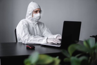 Tıbbi maskeli ve koruyucu elbiseli bir adam, lastik eldivenler evde oturuyor ve karantina sırasında laptopta çalışıyor. Tasarımcı, sanatçı, mimar, iş adamı salgın bir covid 'de uzaktan çalışıyor..