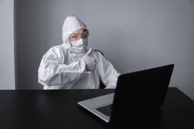 Tıbbi maskeli ve koruyucu elbiseli bir adam, eldivenler evde oturuyor ve karantina sırasında laptopta çalışıyor ve iş ortağına ya da arkadaşlarına baş parmağını gösteriyor. Bir salgın covid içinde uzaktan çalışma.