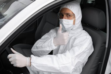 Koruyucu giysili, tıbbi maskeli ve virüs ve bakterilerden korunmak için lastik eldivenli bir adam bir arabada oturuyor ve baş parmağını kaldırıyor. Karantina, dünya salgını, covid 19, koronavirüs, enfeksiyon..