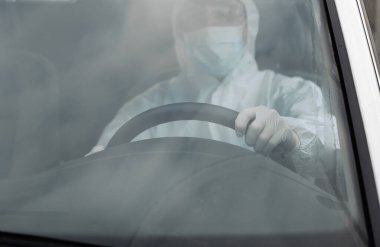 Koruyucu giysili, tıbbi maskeli ve plastik eldivenli bir adam bakteri ve virüsten korunmak için araba kullanıyor. Karantina sırasında koruyucu maske, dünya salgını, covid 19, koronavirüs, enfeksiyon.