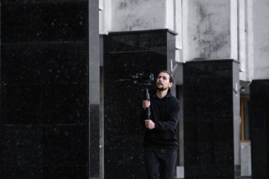 3 eksenli titreşimli dengeleyicide profesyonel kamera tutan genç profesyonel kameraman. Pro ekipmanlar titremeden yüksek kaliteli video çekmeye yardımcı olur. Siyah kapüşonlu bir kameraman video çekiyor..