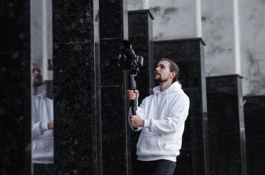 3 eksenli titreşimli dengeleyicide profesyonel kamera tutan genç profesyonel kameraman. Pro ekipmanlar titremeden yüksek kaliteli video çekmeye yardımcı olur. Beyaz kapüşonlu bir kameraman video çekiyor..