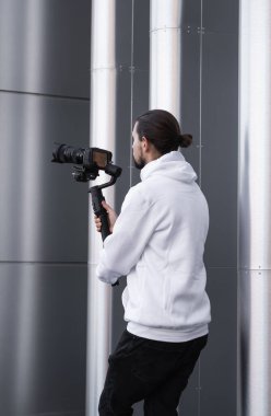3 eksenli titreşimli dengeleyicide profesyonel kamera tutan genç profesyonel kameraman. Pro ekipmanlar titremeden yüksek kaliteli video çekmeye yardımcı olur. Beyaz kapüşonlu bir kameraman video çekiyor..