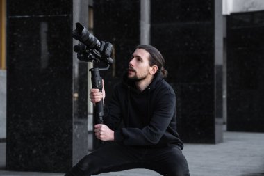 3 eksenli titreşimli dengeleyicide profesyonel kamera tutan genç profesyonel kameraman. Pro ekipmanlar titremeden yüksek kaliteli video çekmeye yardımcı olur. Siyah kapüşonlu bir kameraman video çekiyor..