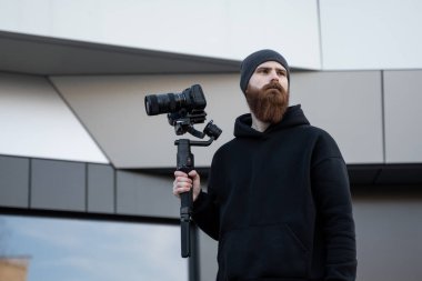 Siyah kapüşonlu sakallı profesyonel kameraman elinde 3 eksenli titreşim dengeleyicisi var. Film yapımcısı profesyonel bir sinema kamerasıyla harika bir video çekiyor. Görüntü Yönetmeni.