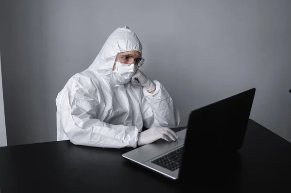Man in medical mask and protective suit, rubber gloves sits at home and works at laptop at the table during quarantine. Designer, artist, architect, businessman at remote work in a pandemic covid. — Stock Photo, Image