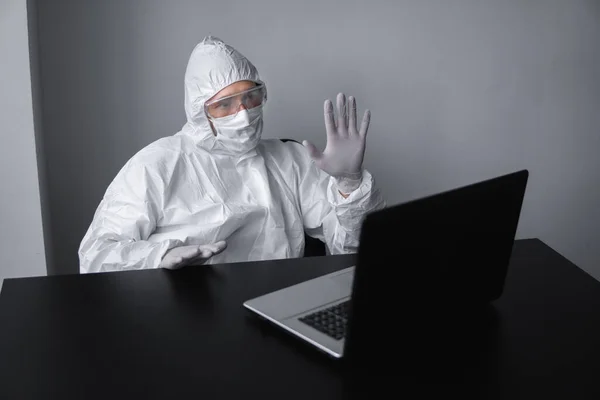 Mann mit medizinischer Maske und Schutzanzug, Handschuhen sitzt zu Hause und arbeitet während der Quarantäne am Laptop am Tisch und grüßt seinen Geschäftspartner oder Freunde. Fernarbeit in einem Pandemiegebiet. — Stockfoto