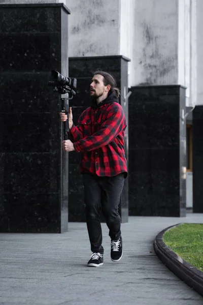 Ung professionell videograf som håller professionell kamera på 3-axlig gimbal stabilisator. Pro-utrustning hjälper till att göra högkvalitativ video utan att skaka. Kameraman bär röd skjorta gör en video. — Stockfoto