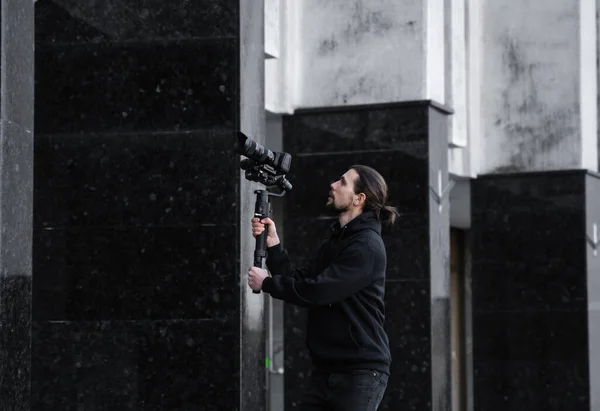 Joven videógrafo profesional con cámara profesional en estabilizador de cardán de 3 ejes. El equipo profesional ayuda a hacer video de alta calidad sin temblar. Camarógrafo con capucha negra haciendo videos . —  Fotos de Stock