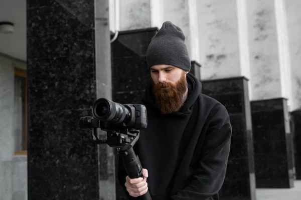 Bearded Professional videographer in black hoodie holding professional camera on 3-axis gimbal stabilizer. Filmmaker making a great video with a professional cinema camera. Cinematographer. — Stock Photo, Image