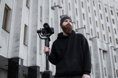 Siyah kapüşonlu sakallı profesyonel kameraman elinde 3 eksenli titreşim dengeleyicisi var. Film yapımcısı profesyonel bir sinema kamerasıyla harika bir video çekiyor. Görüntü Yönetmeni.