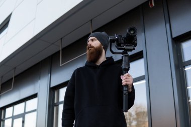 Siyah kapüşonlu sakallı profesyonel kameraman elinde 3 eksenli titreşim dengeleyicisi var. Film yapımcısı profesyonel bir sinema kamerasıyla harika bir video çekiyor. Görüntü Yönetmeni.