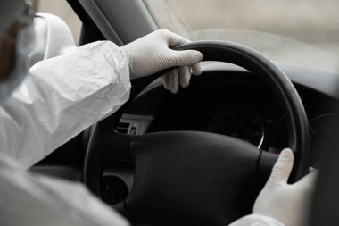 Koruyucu giysili, tıbbi maskeli ve plastik eldivenli bir adam bakteri ve virüsten korunmak için araba kullanıyor. Karantina sırasında koruyucu maske, dünya salgını, covid 19, koronavirüs, enfeksiyon.