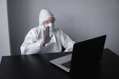 Tıbbi maskeli ve koruyucu elbiseli bir adam, eldivenler evde oturuyor ve karantina sırasında laptopta çalışıyor ve iş ortağına ya da arkadaşlarına merhaba diyor. Bir salgın covid içinde uzaktan çalışma.